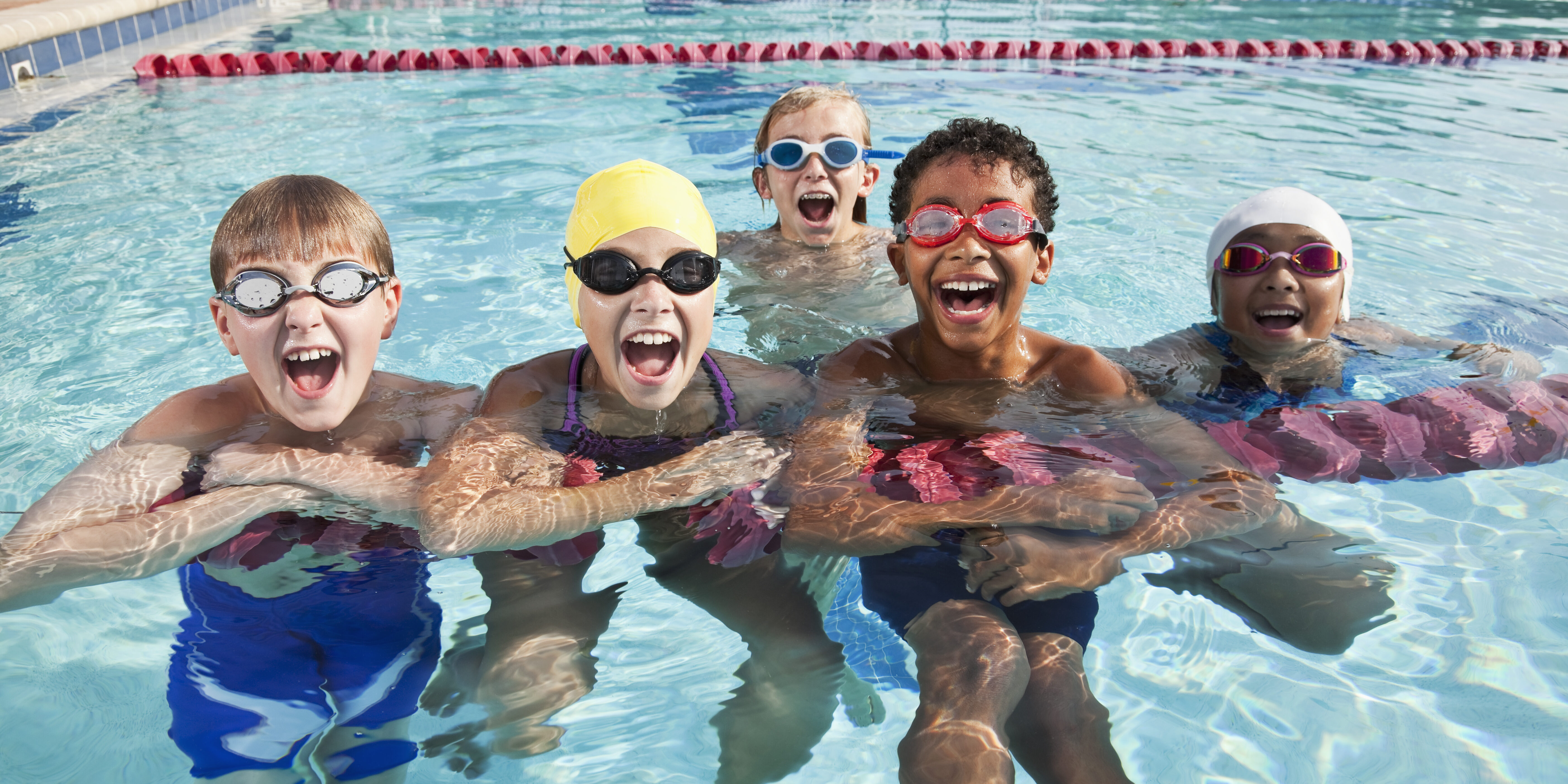 A multiracial group of five children having fun in a swimming pool, hanging onto a lane rope, looking at the camera, laughing and shouting.  The three boys and two girls are 9 to 12 years old.  They are wearing swimsuits and goggles, and the girls have on swim caps.